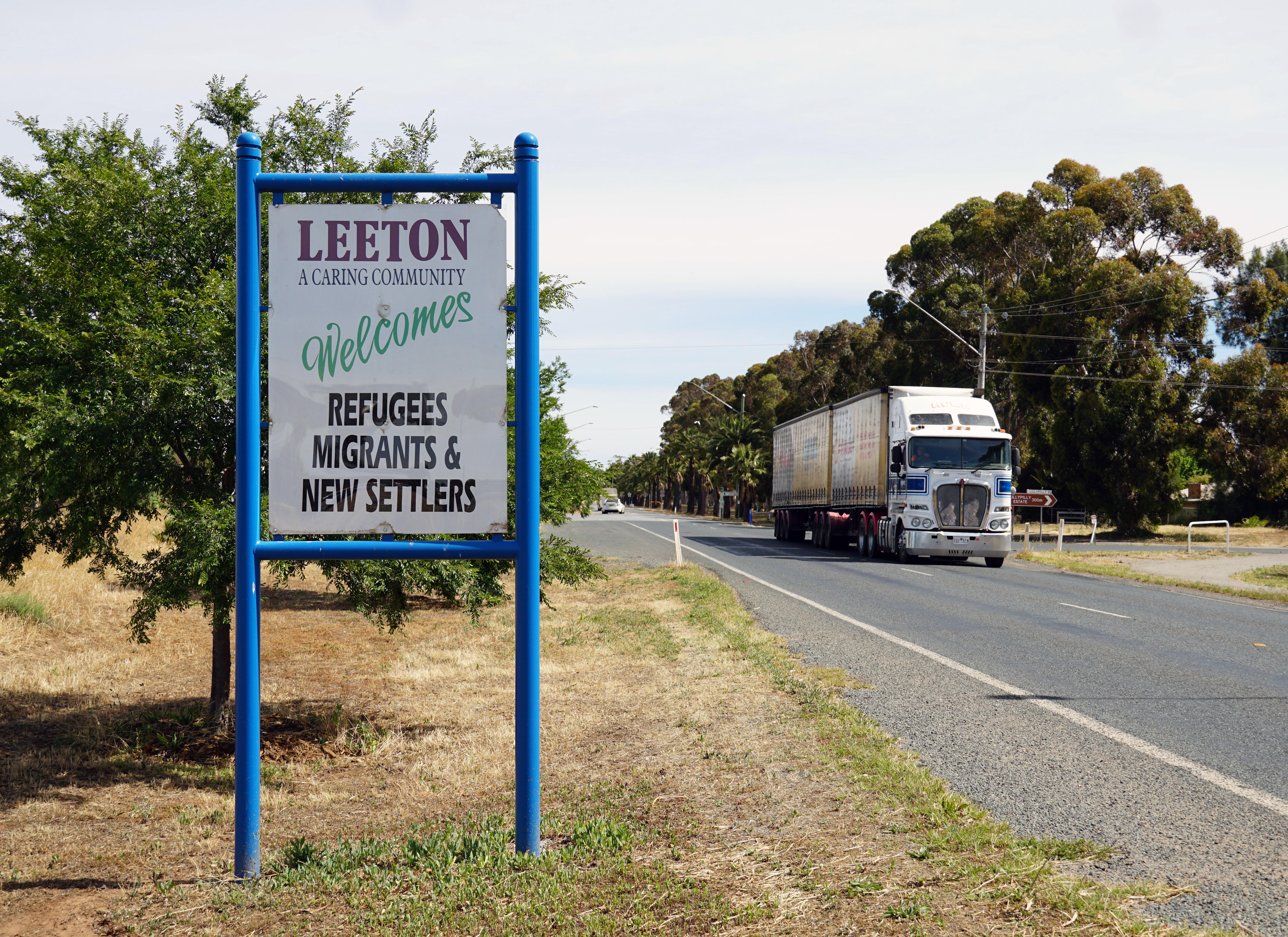 A white sign next to a road reads 'Leeton a caring community welcomes refugees migrants & new settlers', a truck is on the road.