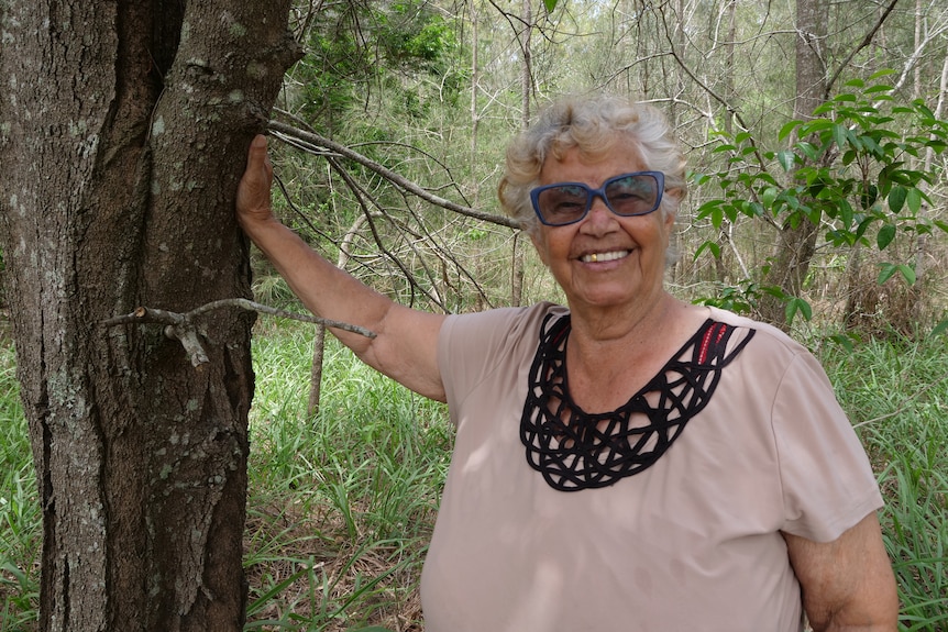 An elderly Aboriginal woman with her hand on a tree smiling in the bush.