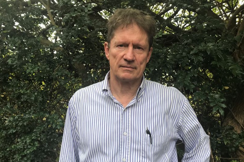 A man with short hair, wearing a business shirt, standing in front of some trees.