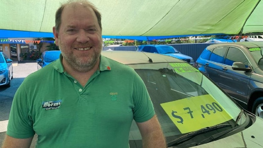 A man with a green shirt stands in front of a car for sale. He is smiling.