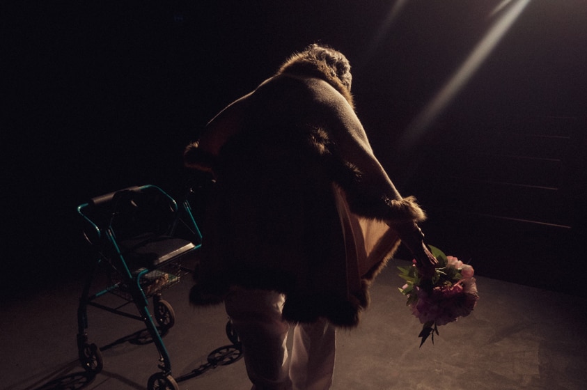 An older woman stands with her back to camera and takes a bow holding on to her walker.