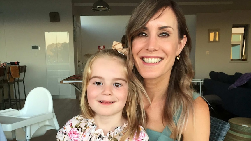 A woman holds her young daughter on her knee, both smiling at the camera.