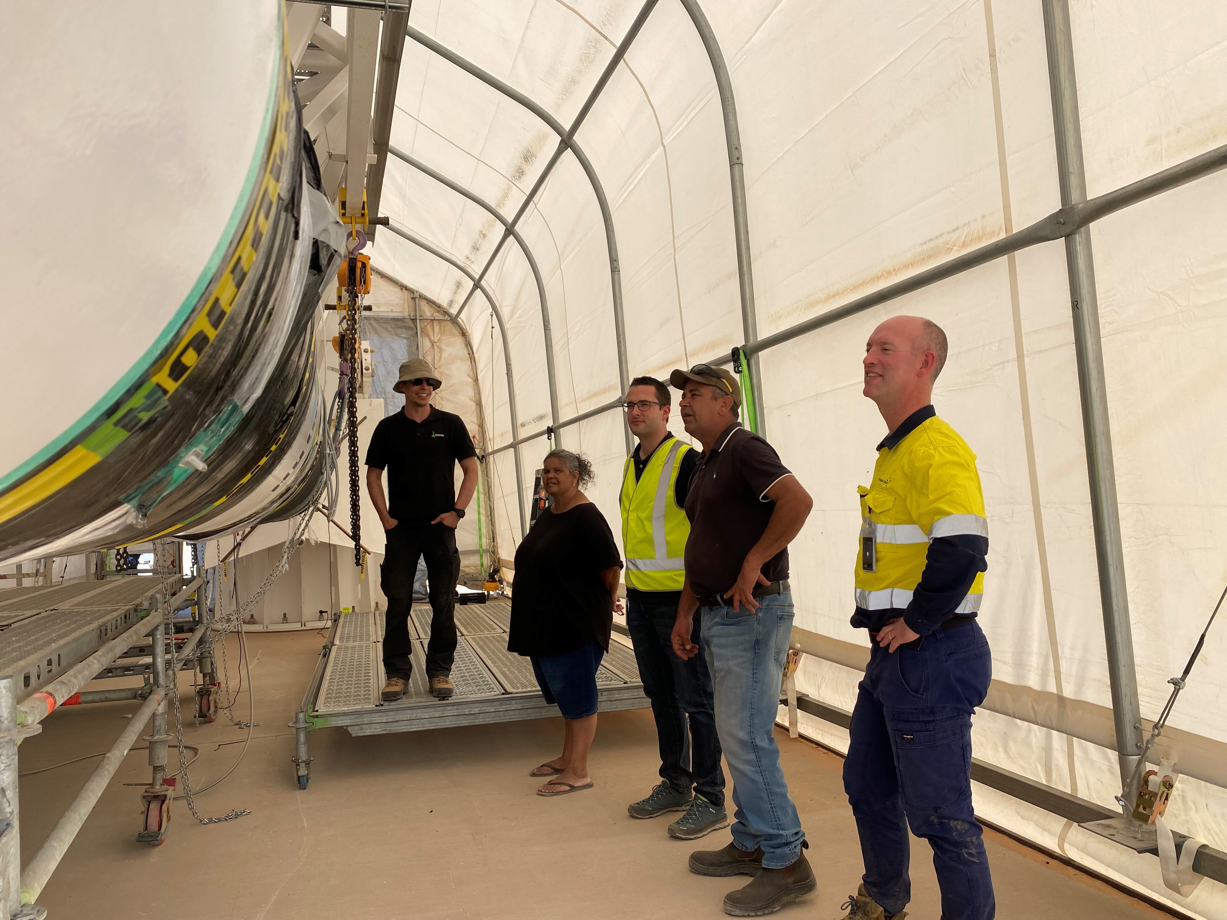 Five people standing on right in large white tent, looking rocket lying on its side, rocket obscured 