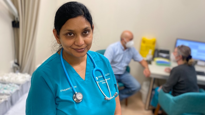  Dr Sonu Haikerwal stands in a treatment room at her practice, with a patient and nurse sitting behind her