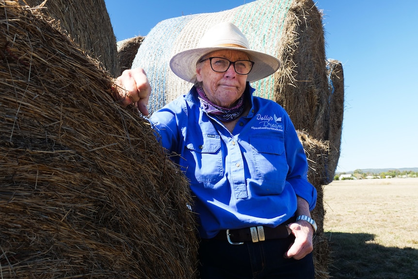 A woman in a blue shirt and wide brim hat leans on hay bales and looks into the camera. 