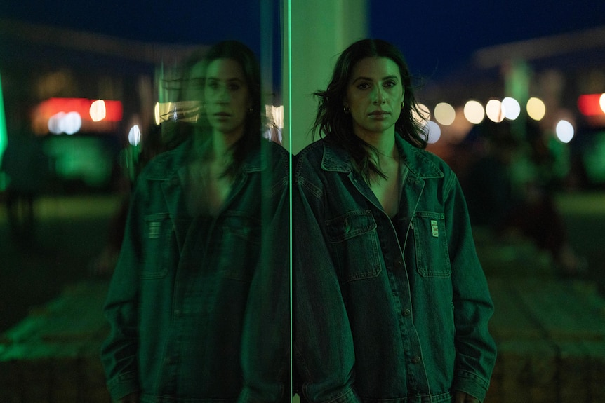 Woman in a denim jacket leaning against a glass window at night.