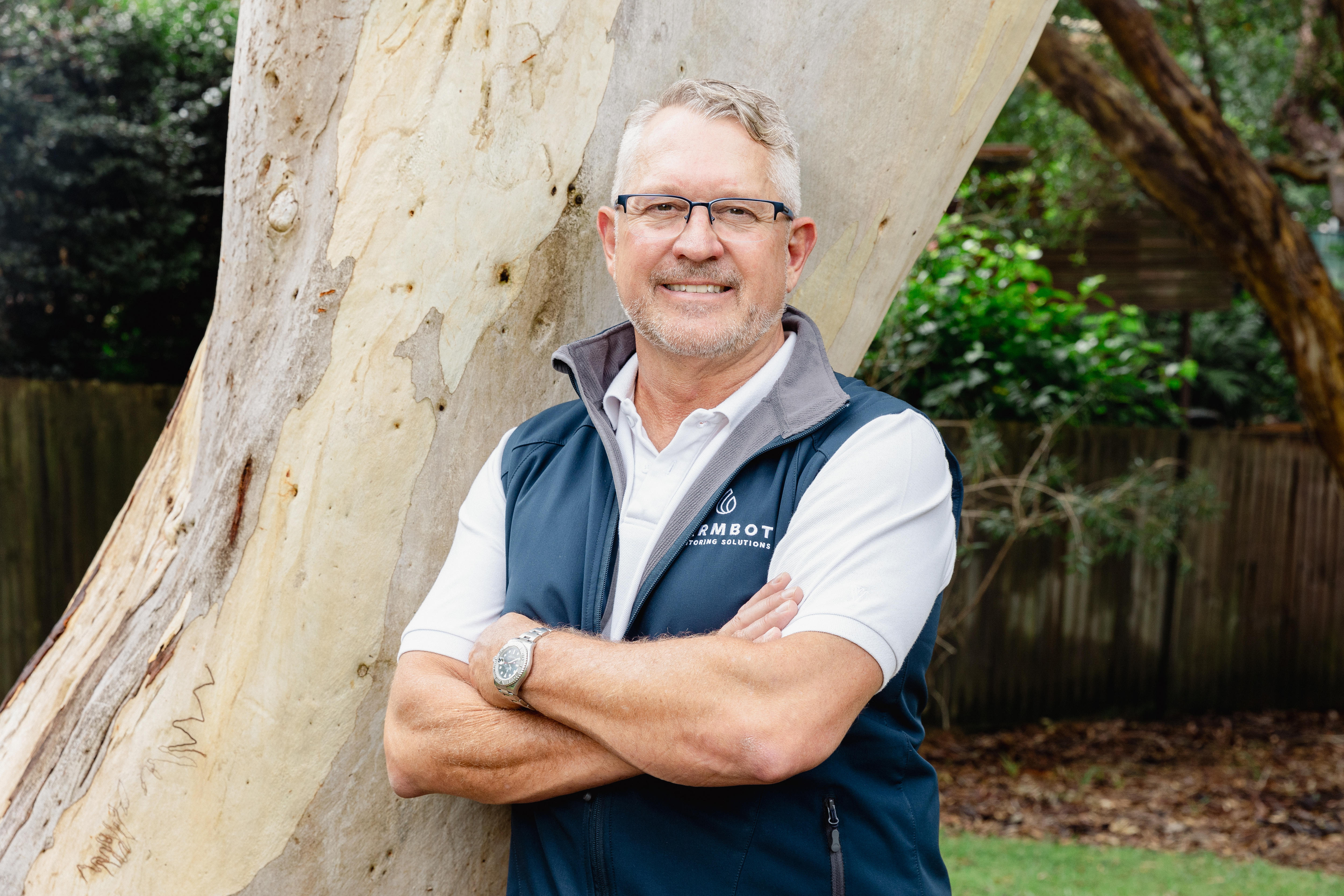 A smiling, silver-haired man leans against a tree.