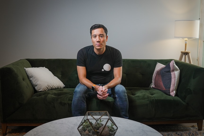 A man sits on a green velvet couch, wearing a black tshirt and jeans, staring into the camera.