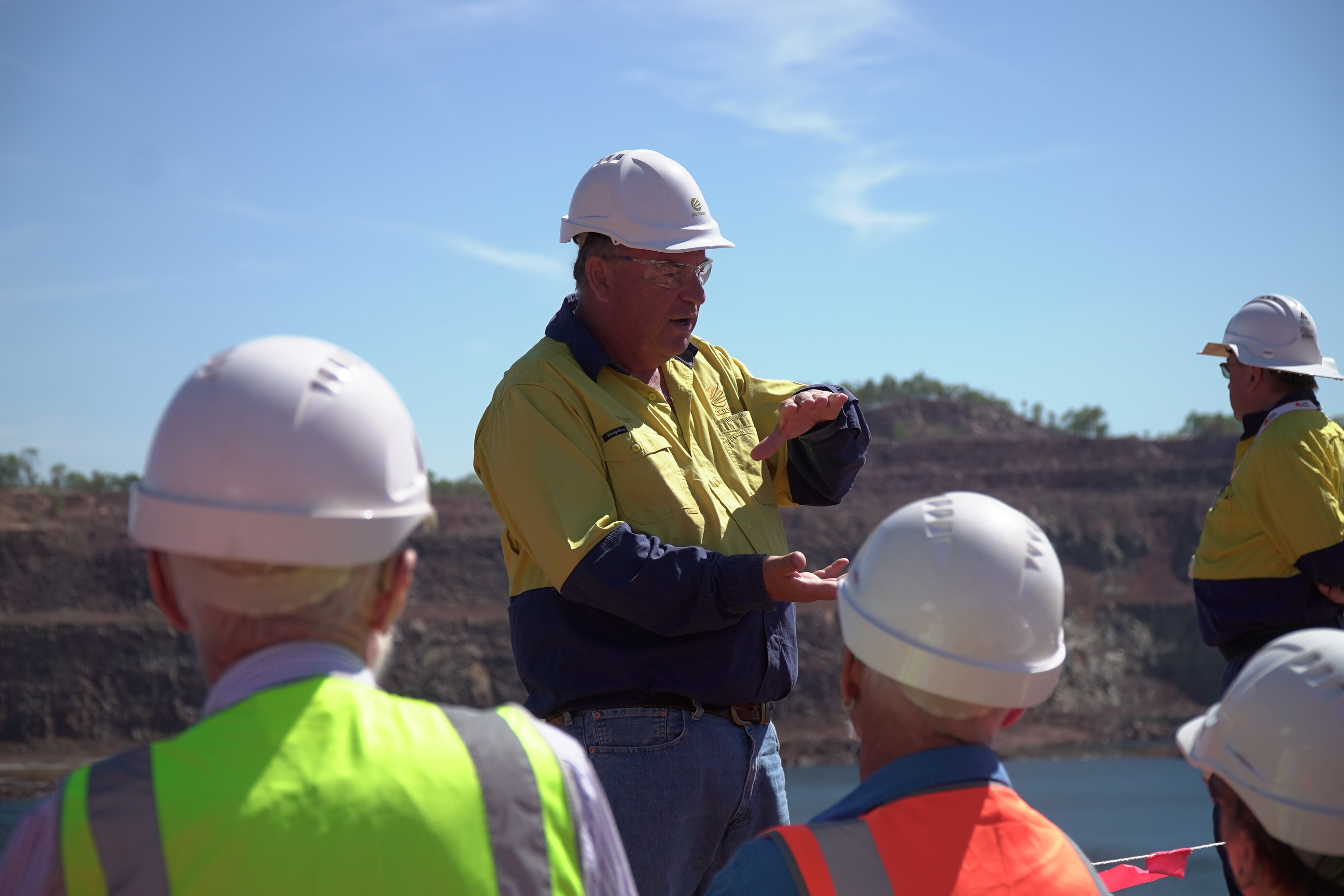 A man in a white hard hat and high vis yellow shirt gestures to a group of people seen in the foreground all wearing hard hats