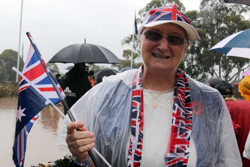 A lady with a Union Jack hat, scarf and flags smiles in a rainy Canberra.