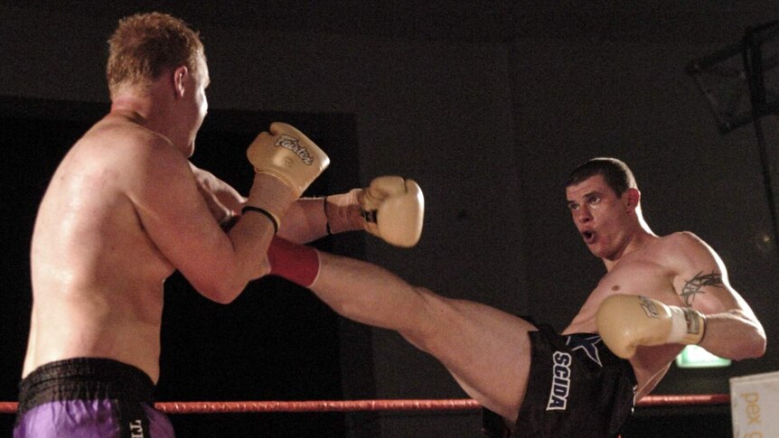 Two men on a boxing ring fighting, with one kicking another in a long extended kick.