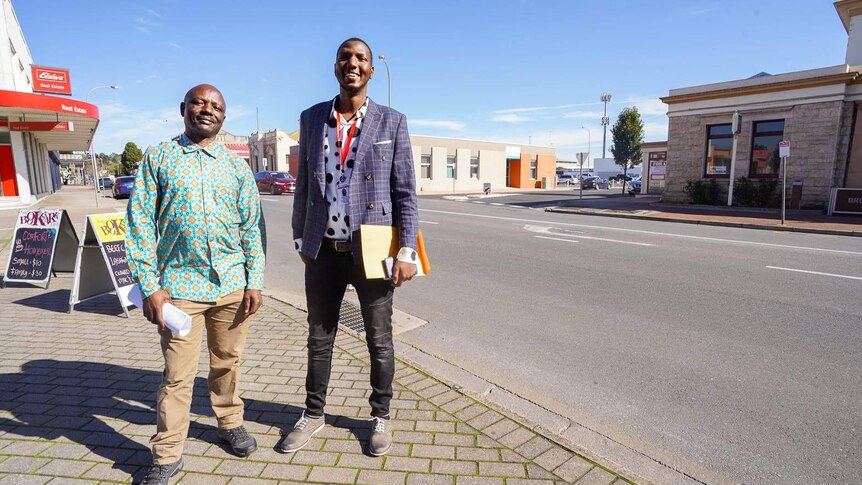 A shorter man in a bright patterned shirt smiles next to a tall man in a suit jacket next to Mount Gambier's main street.