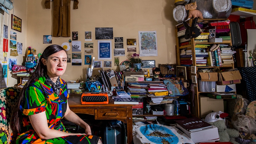 The writer, wearing a brightly printed day dress,  sits with her dayglo orange typewriter at a cluttered desk.