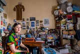 The writer, wearing a brightly printed day dress,  sits with her dayglo orange typewriter at a cluttered desk.