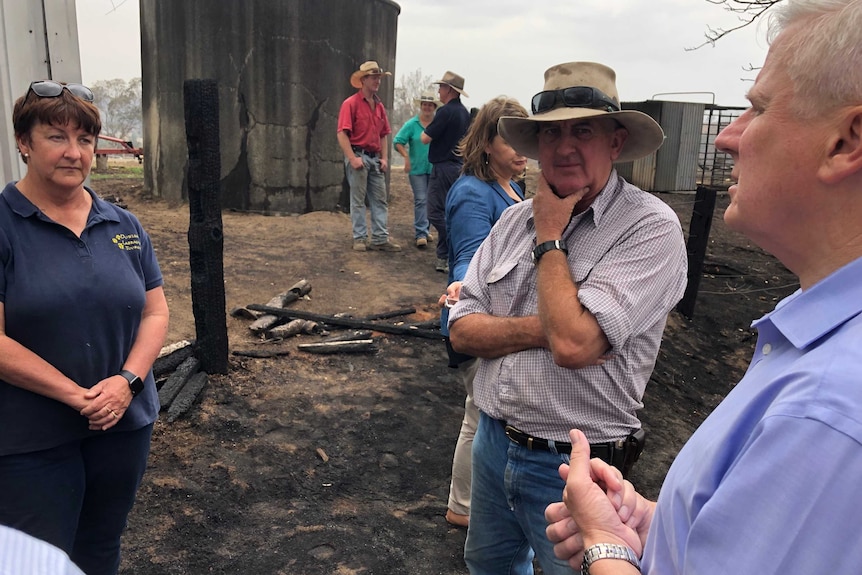 A politician speaks to farmers standing near burnt-out rubble