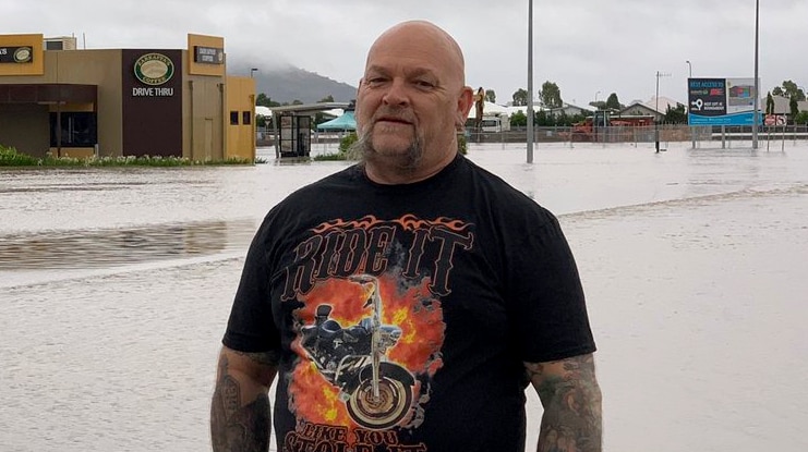 Male truck driver standing in front of flood waters, wearing black clothing, tattoos but a small smile.
