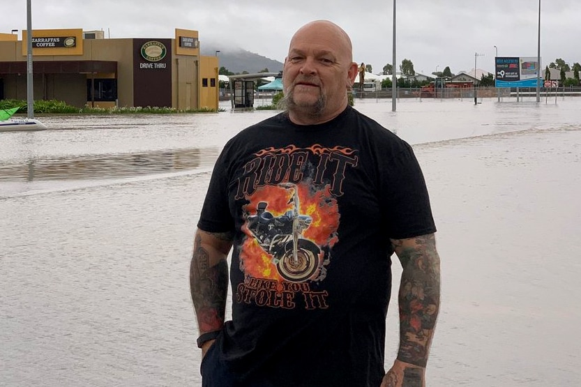 Male truck driver standing in front of flood waters, wearing black clothing, tattoos but a small smile.