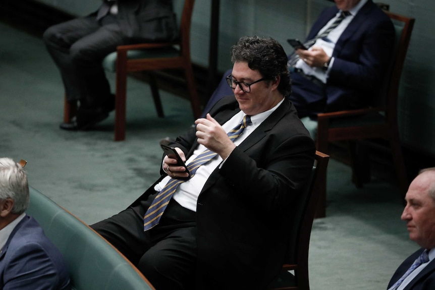 Christensen sits in his chair at the back of the chamber holding and looking at his phone and smirking.