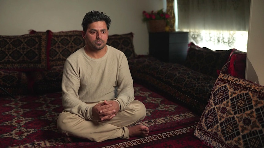 A man wearing a beige top and pants sits on a rug.