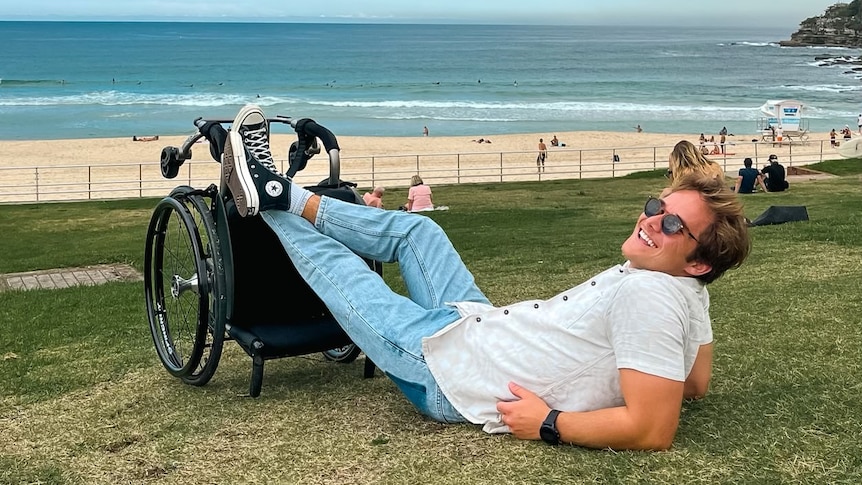 An image of Jimmy laying on the grass with his legs propped up on his wheelchair the beach in the background