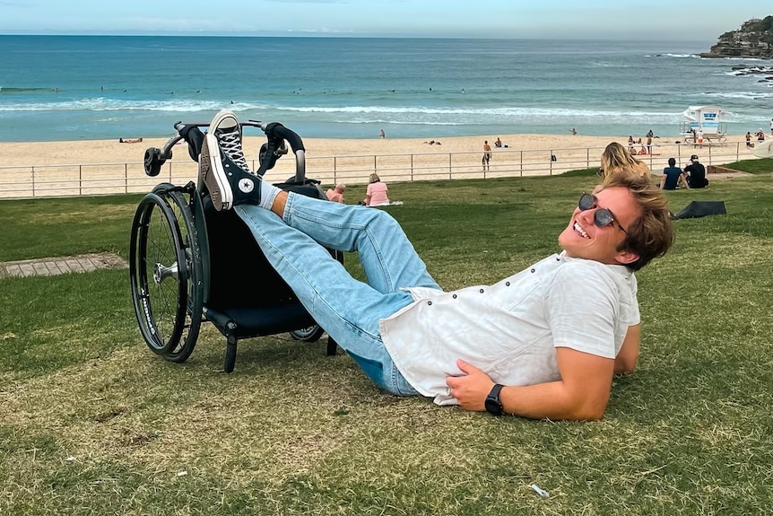 An image of Jimmy laying on the grass with his legs propped up on his wheelchair the beach in the background