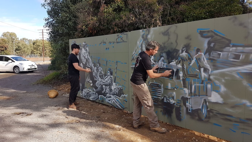 Two men hold spray cans and create art on a green wall.