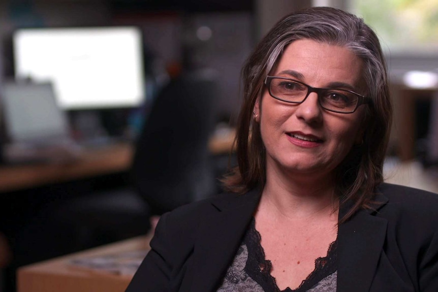 MCU of Jo Natoli sitting in her office wearing black rimmed glasses, black jacket and black and grey shirt