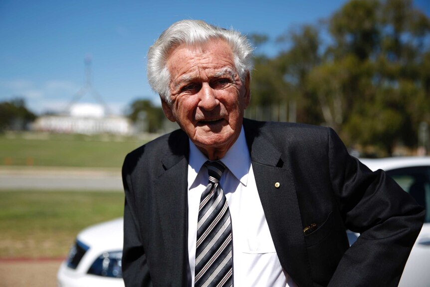 Bob Hawke, wearing a black suit and diagonally striped tie, looks into the sun. Parliament House is visible in the background.