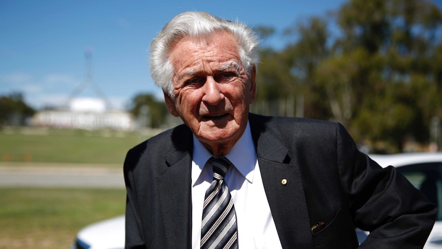 Bob Hawke, wearing a black suit and diagonally striped tie, looks into the sun. Parliament House is visible in the background.