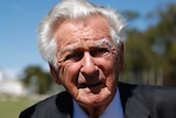 Bob Hawke, wearing a black suit and diagonally striped tie, looks into the sun. Parliament House is visible in the background.