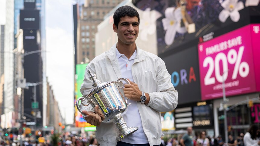 A tennis player wearing a cream jacket, white shirt and jeans stands in the middle of New York holding a trophy.