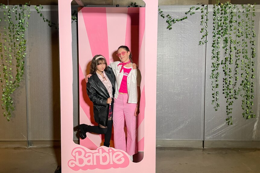 Two teenagers smile in pink Barbie-inspired outfits, in a photobooth set up for the occasion.