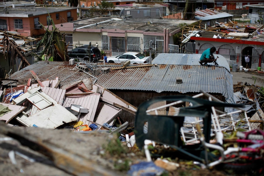 A man, surrounded by damaged houses, tries to fix a roof.