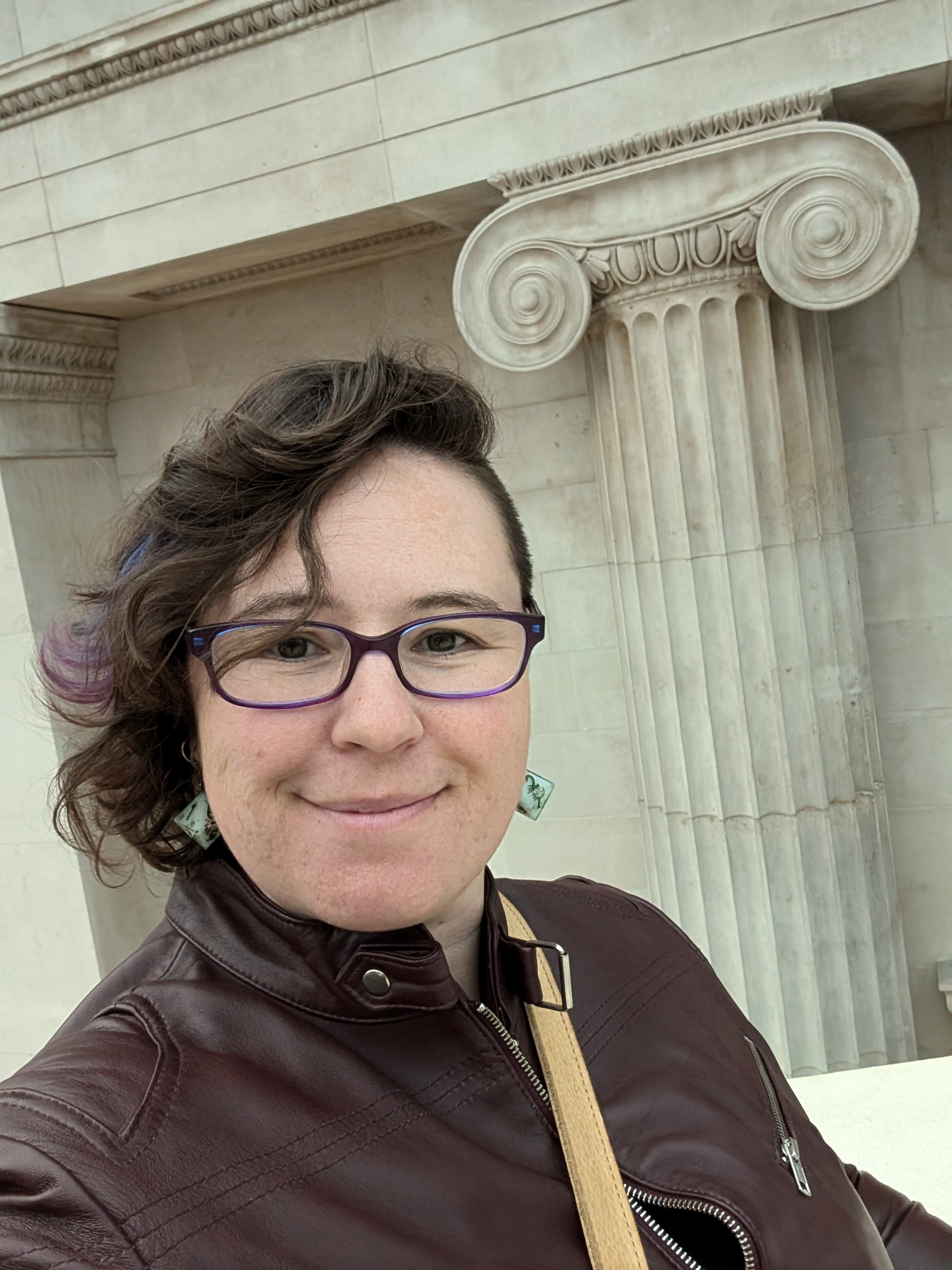 A woman with dark brown hair, glasses and a brown jacket in front a building with a classical-style pillar.