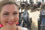 A smiling blonde woman showing her teeth in front of a row of motorbikes in Bali.