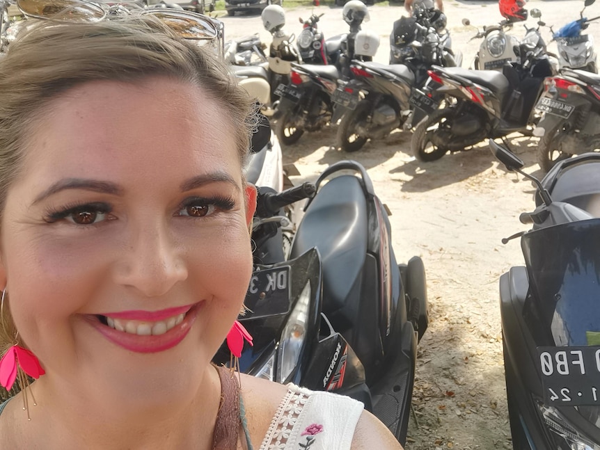 A smiling blonde woman showing her teeth in front of a row of motorbikes in Bali.