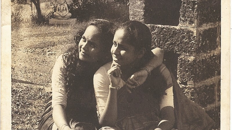 A sepia toned photo of two women sitting in saris looking away from the camera, one has her arm around the other.