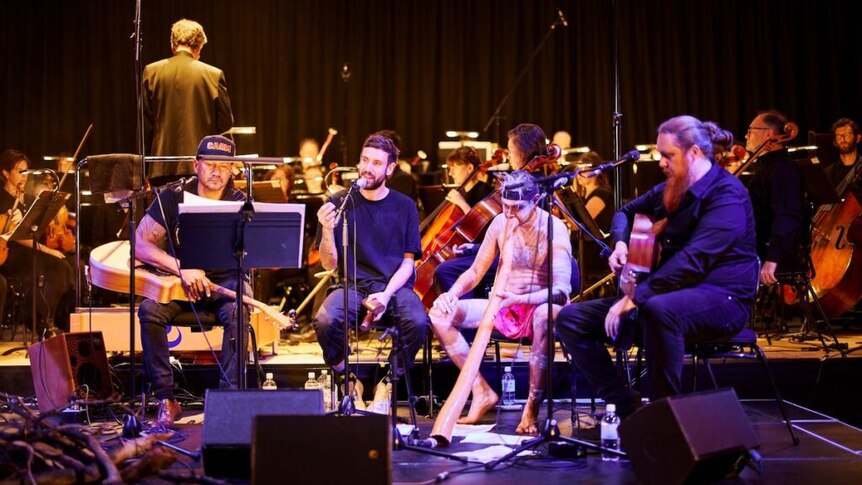 Four soloists sit in front of the Adelaide Symphony Orchestra onstage, one of them an indigenous yidaki performer.