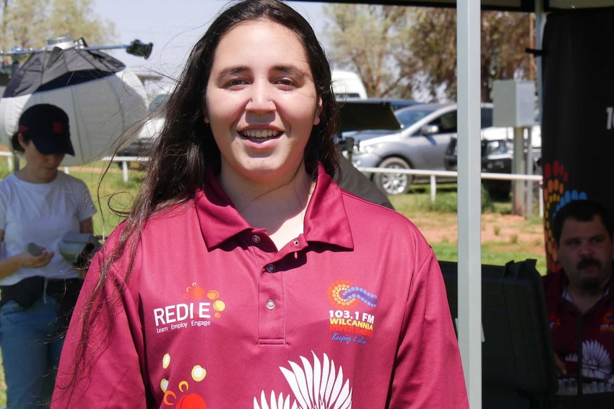 A young woman in a maroon t-shirt smiles at the camera standing in front of a Wilcannia River Radio-branded marquee.