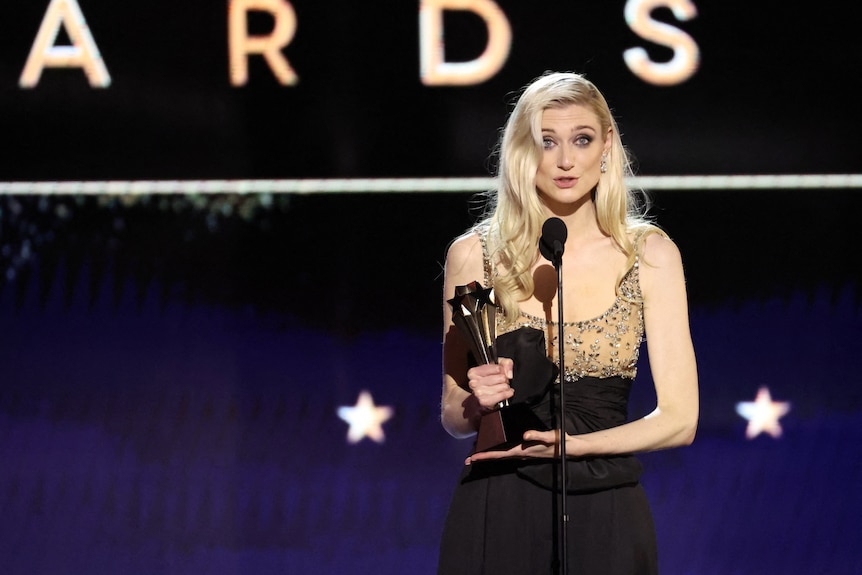 Elizabeth Debicki holding an award, long blonde hair, gold evening gown, talking into a microphone