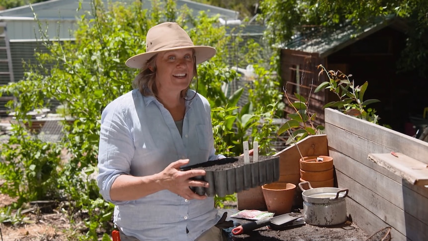 Millie Ross stands in her garden holding a seedling tray. She wears a tan hat and a light shirt and it's a sunny day.