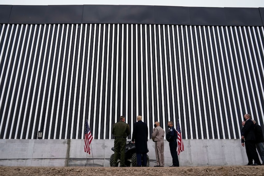 A large high sectionsof wall with some people at the bottom of it, including Donald Trump