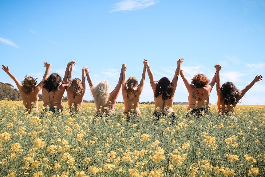 Eight women wearing black underwear stand with their arms up in the air, looking over a canola field