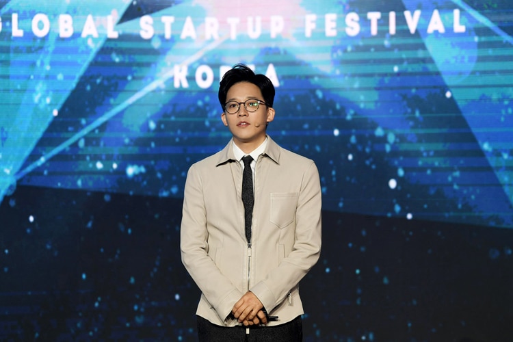 A young man wearing a beige jacket over white shirt and black tie stands in front of a screen