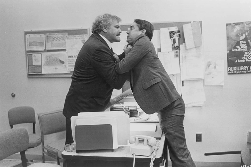A black and white photo of Dennehy grabbing a man by the shirt over a desk.