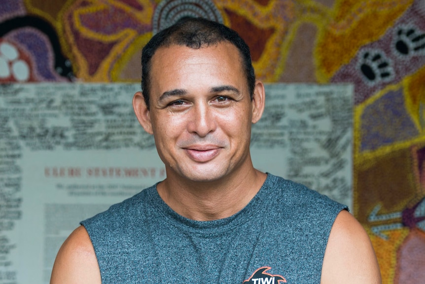 A man in a singlet smiles at the camera, an Indigenous artwork visible behind him.