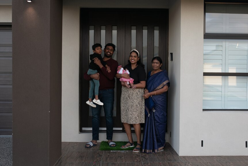 A man holds a boy, woman holds baby, stand next to woman in blue silk saree in front of house. All smiling.
