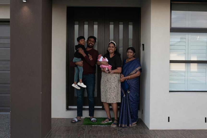 A man holds a boy, woman holds baby, stand next to woman in blue silk saree in front of house. All smiling.