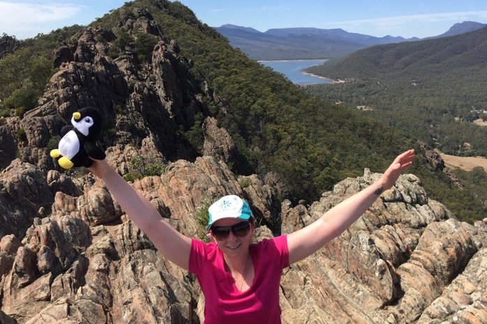 Kylie holds her arms up with joy on the top of a mountain, a view behind her. She holds a toy penguin.