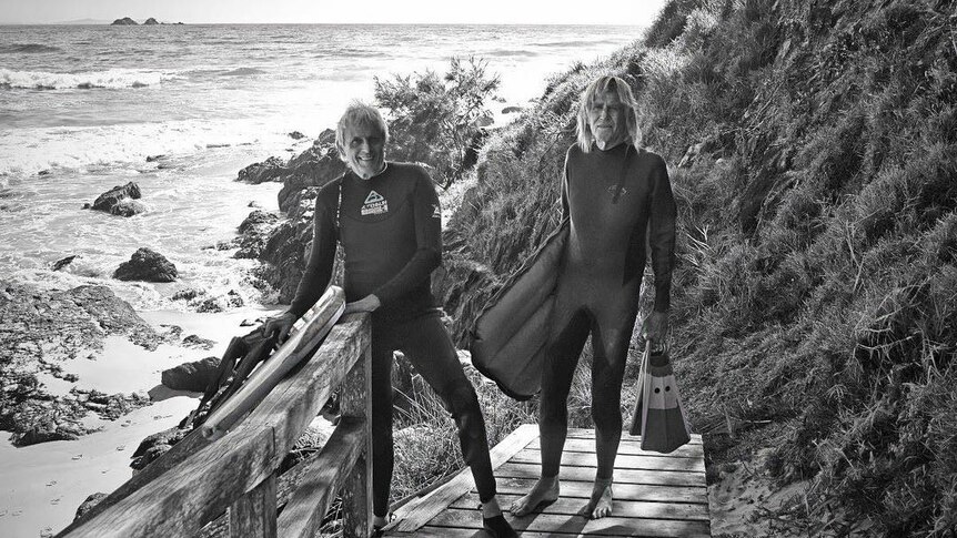 Two men in wetsuits stand on the path down to the beach, holding their surf gear.